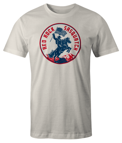 White Squatchrider t-shirt with red text. Features Sasquatch riding a horse and waving an American flag. A patriotic and unique design.